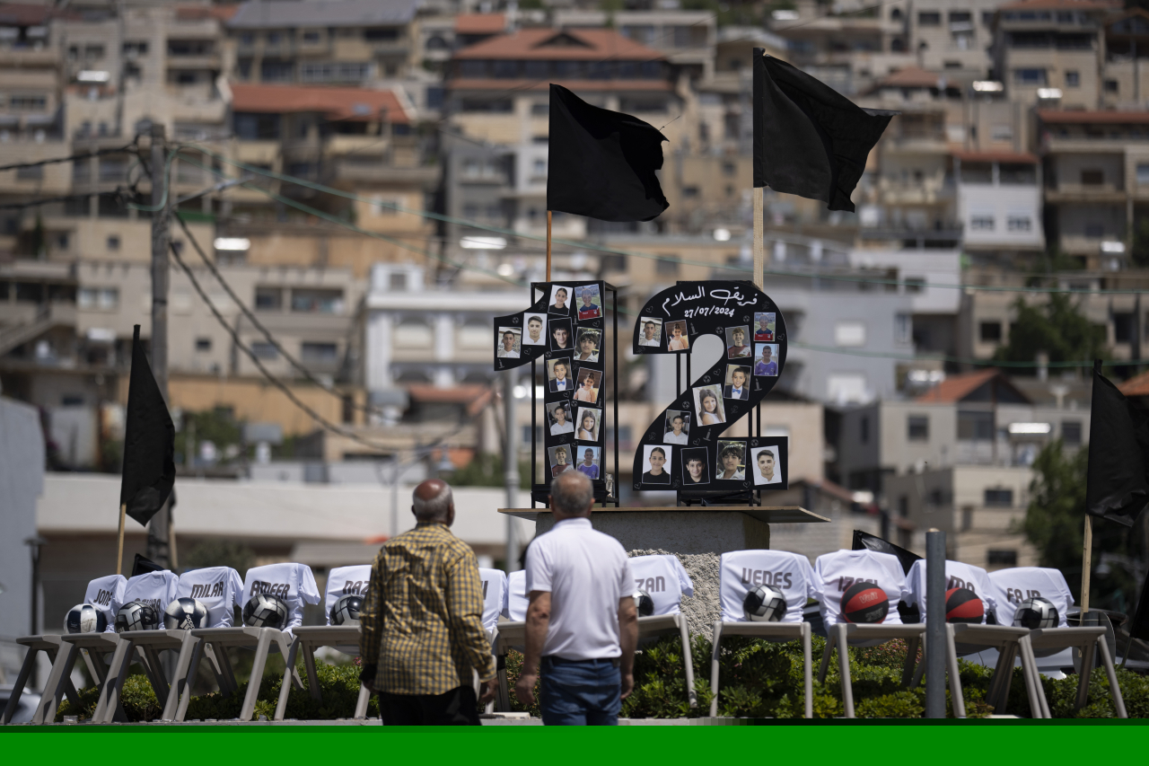 People stand in front photos of the 12 children and teens, killed in a rocket strike at a soccer field, and chairs with their names written on it, displayed on a roundabout in the village of Majdal Shams, in the Israeli-annexed Golan Heights, Tuesday. (AP-Yonhap)